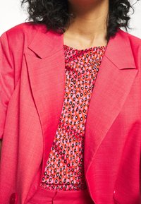 Růžový blejzr s klopami přes vzorovaný top s růžovými, oranžovými a černými skvrnami. Texturovaná tkanina s knoflíkovým detailem.