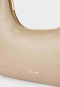 Μπεζ υφασμένη δερμάτινη τσάντα ώμου με χρυστό φερμουάρ και μικρό χρυσό λογότυπο Calvin Klein στο μπροστινό μέρος.