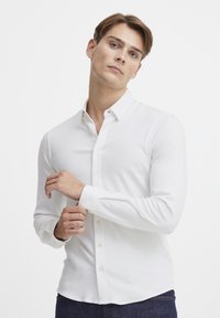 Pitkähihaisessa valkoisessa paidassa on nappikaulus, sileä pinta ja istuva muotoilu. Paidassa on napit edessä ja hihansuiden kohdalla.