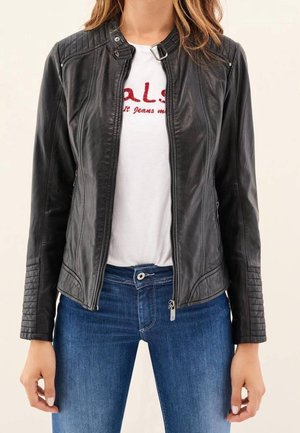 Nainen pukeutuneena istuvaan mustaan nahkatakkiin valkoisen graafisen t-paidan ja polvipituisten sinisten farkkujen kanssa, seisomassa neutraalia taustaa vasten.