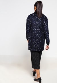 Donna che si allontana indossando un cappotto leopardato blu scuro, pantaloni culotte neri a righe e scarpe con tacco blocco nere su uno sfondo bianco.