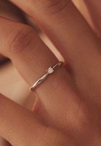 Zilveren ring met een gladde band, voorzien van een kleine ronde diamant, gezet in een zes-punts montuur, gedragen op een vinger tegen een gestructureerde achtergrond.