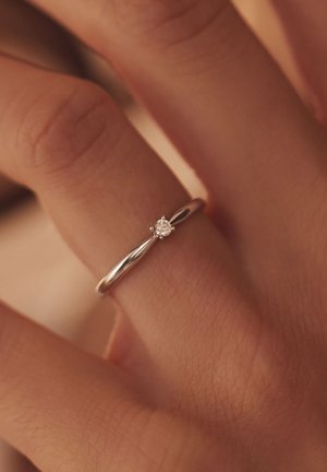 Silberner Ring mit glatter Fassung, der einen kleinen runden Diamanten in einer sechszackigen Fassung zeigt, der an einem Finger vor einem strukturierten Hintergrund getragen wird.