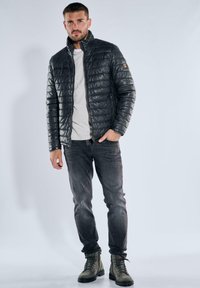 Schwarze gesteppte Jacke mit hohem Kragen und Reißverschluss, getragen über einem hellgrauen Hemd. Kombiniert mit dunkelblauer Jeans und grünen Stiefeln.