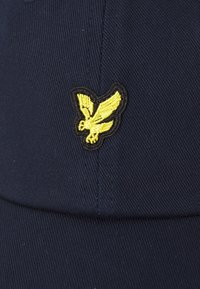 Mørkeblå caps laget av teksturert stoff, med en brodert ørn i gult og svart på forsiden.