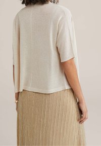 Světlý béžový svetr s krátkými rukávy, volným střihem a mírně zkráceným lemem. Nošený přes plisovanou zlatou metalickou sukni.