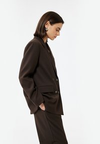 Bruine blazer met een dubbelgebreid ontwerp, met gesneden revers en twee voorzakken. Gelaagd stof met een subtiele glans, gecombineerd met bijpassende broek.