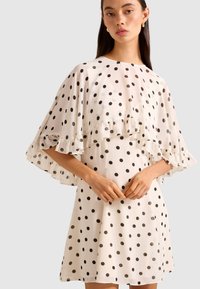 Woman wearing a white dress with black polka dots, featuring a loose ruffled cape overlay and gold hoop earrings.