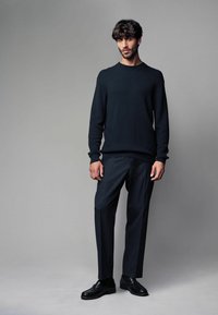 Dunkel gemusterter Pullover, gerippte Bündchen und dunkel gestreifte Hose, kombiniert mit glänzenden schwarzen Loafers. Vor einem neutralen Hintergrund.