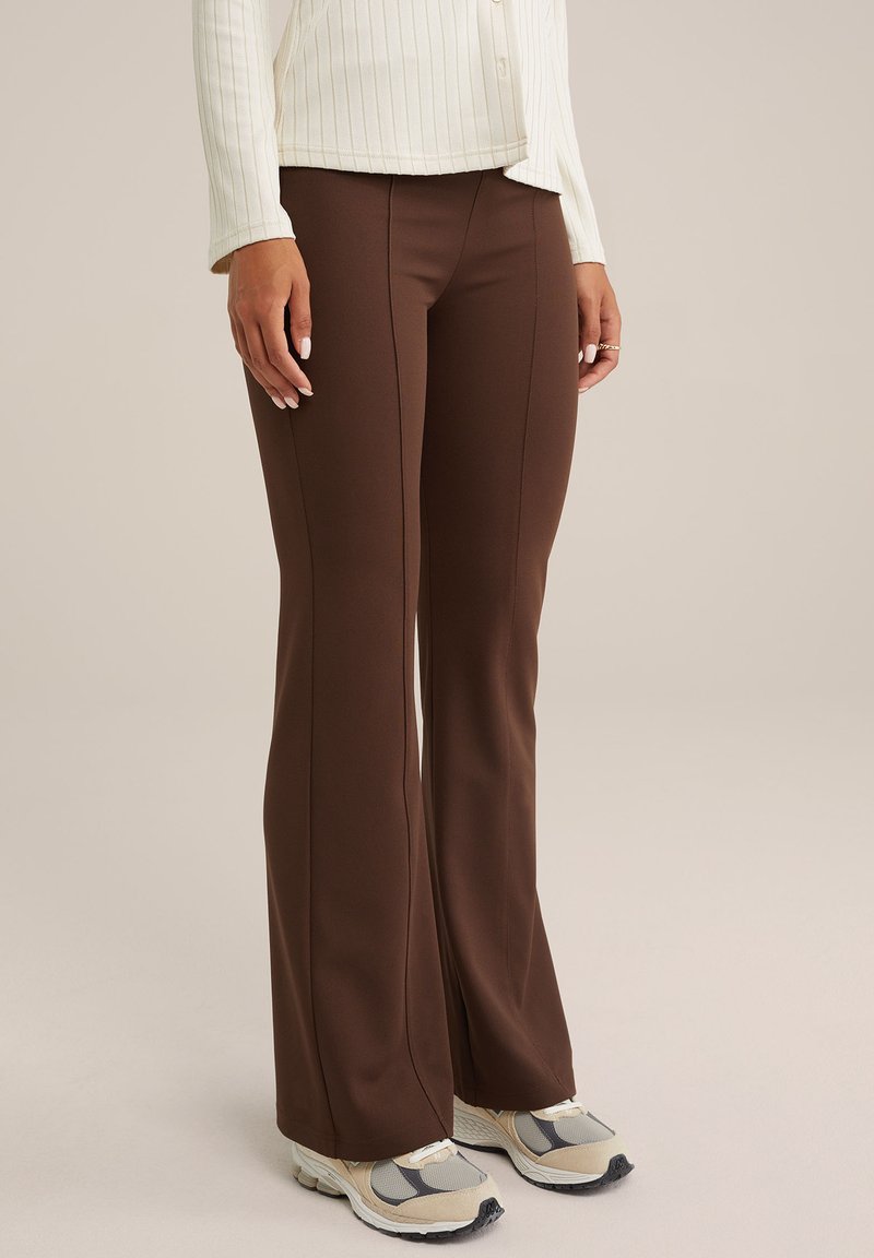 Pantalones marrones acampanados de tela elástica, con una textura suave y detalles en las costuras laterales. Combinados con zapatillas deportivas grises y beige.