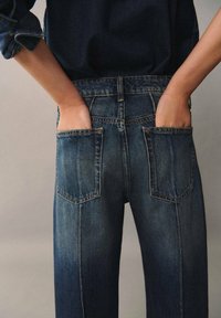 Jeans de mezclilla azul oscuro con cintura alta, que cuentan con dos bolsillos traseros, un diseño de piernas rectas y un ligero desgate en los muslos.