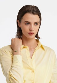 Bratara de aur cu o piatră mică, de culoare verde deschis, purtată pe un încheietură goală. Modelul poartă o cămașă pale galbenă cu nasturi.