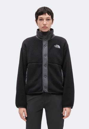 Persona con cabello corto y oscuro lleva una chaqueta de forro polar negra y pantalones negros, ambos con la marca The North Face, de pie contra un fondo claro y liso.