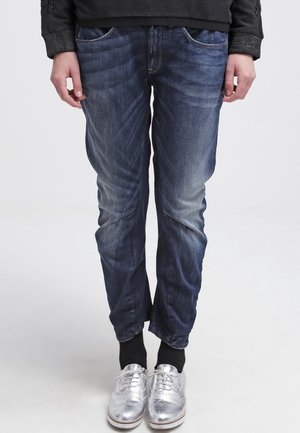 Person iført falmede blå jeans, sorte sokker, metalsølvfarvede wingtip-sko og en sort langærmet top, stående mod en ensfarvet baggrund.