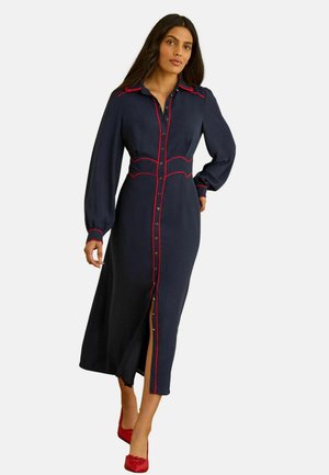 Kobieta ubrana w granatową, midi sukienkę zapinaną na guziki z długim rękawem, ozdobioną czerwonymi lamówkami, oraz czerwone buty na wysokim obcasie, idąca do przodu.