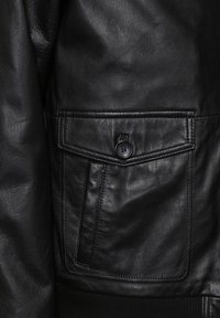 Zwarte leren jas met een enkele knoopsluiting, een klepzak en geribbelde gebreide accenten aan de zoom. Soepele textuur en matte afwerking.