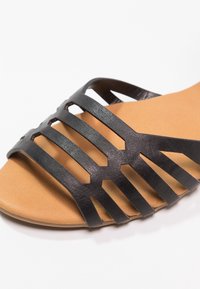 Black strappy sandal featuring cut-out details, a smooth, matte finish, and a tan cushioned insole. Predominantly flat sole design.