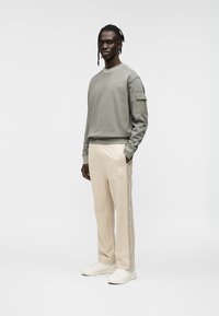 Γκρι βαμβακερό φούτερ με μακρύ μανίκι και τσέπη. Μπεζ παντελόνι χαλαρής εφαρμογής με λογότυπο στο πλάι. Λευκά αθλητικά παπούτσια.