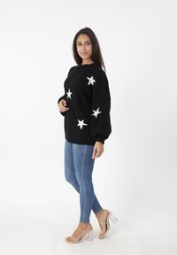 Zwarte gebreide trui met witte sterpatronen, gecombineerd met blauwe skinny jeans en transparante hakken. Soepele, gestructureerde stof.