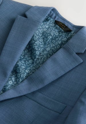 Gaiši zils žakete ar faktūru virsmu un ziedu raksta oderi iekšpusē. Raksturo ar izspraustiem atlokiem un vienu krūšu kabatu.