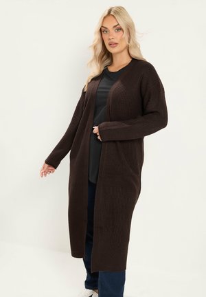 Donna bionda che indossa un lungo cardigan marrone scuro lavorato a maglia sopra una maglietta nera, jeans blu e scarpe bianche, in piedi davanti a uno sfondo semplice.