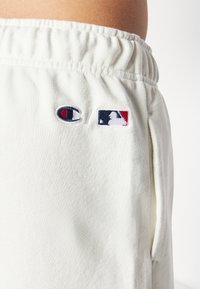 Baltas kokvilnas šorts ar elastīgu jostasvīru, kuram ir izšūtas Champion un MLB logo kreisajā pusē un sānu kabatas.