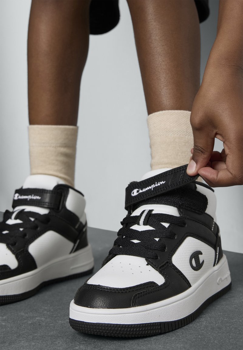Personne ajustant la basket montante Champion noir et blanc avec des chaussettes beige et des lacets noirs sur un sol gris.
