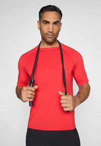 Κόκκινη αθλητική μπλούζα κοντομάνικη με στρογγυλή λαιμόκοψη, απαλή υφή, φορεμένη από άτομο που κρατάει ένα σχοινάκι, ορατά μαύρα σορτς.