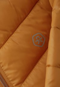 Orange fluffig jacka i tyg med en slät textur, som har sömda sektioner och en hexagonal logotyp i grått. Dragkedjedetalj synlig på sidan.