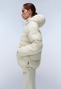 Zachte beige pufferjack met een capuchon, voorzien van een ritssluiting, zijzakken en elastische manchetten. Zachte, gladde textuur. Eenvoudig, modern ontwerp.