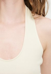 Κοντινό πλάνο ενός ατόμου που φοράει ριμπδωτό εκρού τοπ με halter, δείχνοντας λαιμό, ώμο και άνω στήθος με χαλαρές τούφες μαλλιών.