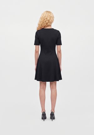 Femme aux longs cheveux blonds bouclés, vêtue d'une courte robe noire et de chaussures à talons noirs, debout dos à la caméra.