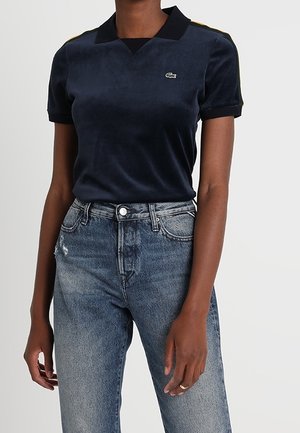 Osoba ubrana w granatową koszulę z krótkim rękawem, czarnym kołnierzykiem i logo, zestawioną z wysokim stanem jasnoniebieskich, wyblakłych dżinsów.