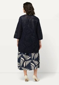 Cárdigan de ganchillo azul marino con detalles brillantes, combinado con una falda azul marino que presenta un patrón de hojas en color crema. La modelo lleva zapatos de tacón claros.