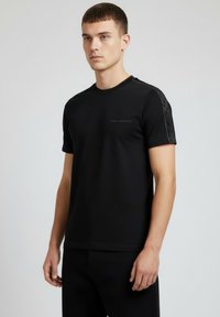 Czarny t-shirt z krótkim rękawem o fakturowanym wzorze na ramionach, z subtelnym nadrukiem logo na piersi. Gładka tkanina, dekolt w serek.