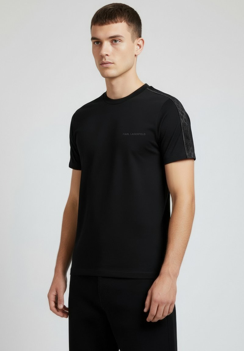 Czarny t-shirt z krótkim rękawem o fakturowanym wzorze na ramionach, z subtelnym nadrukiem logo na piersi. Gładka tkanina, dekolt w serek.