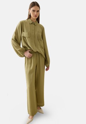 Femme portant une chemise ample à manches longues vert olive avec des poches boutonnées et un pantalon large assorti, debout dans des chaussures beiges pointues.