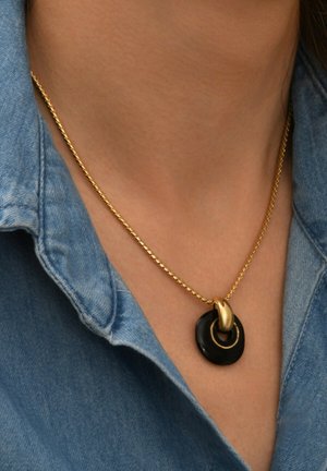Collier en chaîne dorée avec un pendentif circulaire noir et un petit détail en forme de cercle doré. Porté sur une chemise en denim bleu clair.