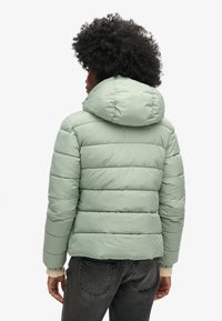 Giacca imbottita in morbido verde menta con cuciture orizzontali e ampio cappuccio attaccato. Presenta polsini elasticizzati e contrasto di texture sull'orlo.