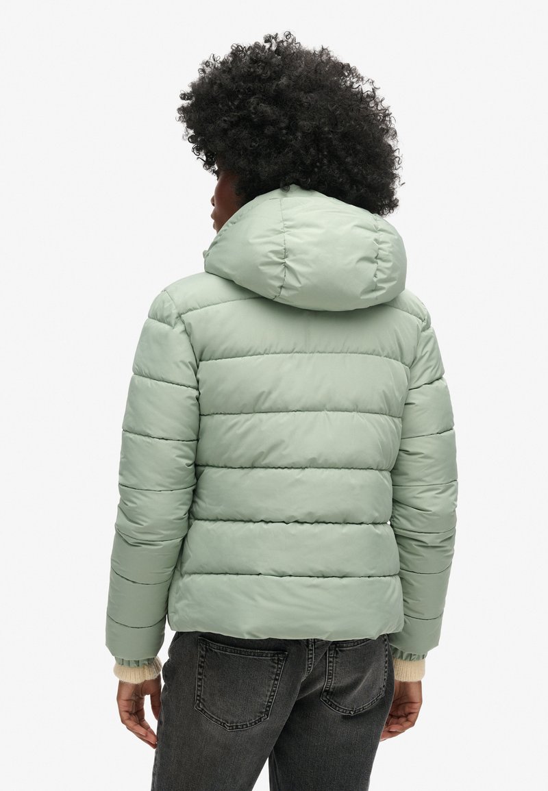 Jasnozielona kurtka puchowa z poziomymi przeszyciami, kapturem i ściągaczami na rękawach. Widok z tyłu ukazuje gładkie, matowe wykończenie oraz zorganizowaną konstrukcję.