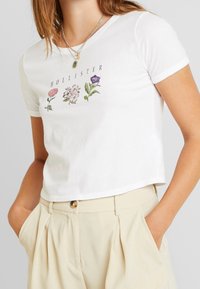 White cotton cropped t-shirt with floral embroidery in pink, purple, and green. Crew neck and short sleeves. Paired with beige trousers.