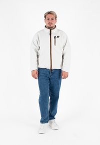 Biała polarowa kurtka z kołnierzem stójkowym, zapięciem na zamek z przodu i pomarańczowym paskiem akcentującym; noszona na niebieskich dżinsach i białych trampkach.