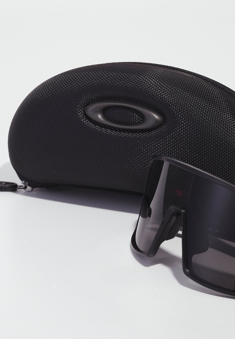 Lunettes de soleil noires avec une forme de lentille rectangulaire, dotées d'une monture mate, placées à côté d'un étui rigide noir texturé avec un logo.