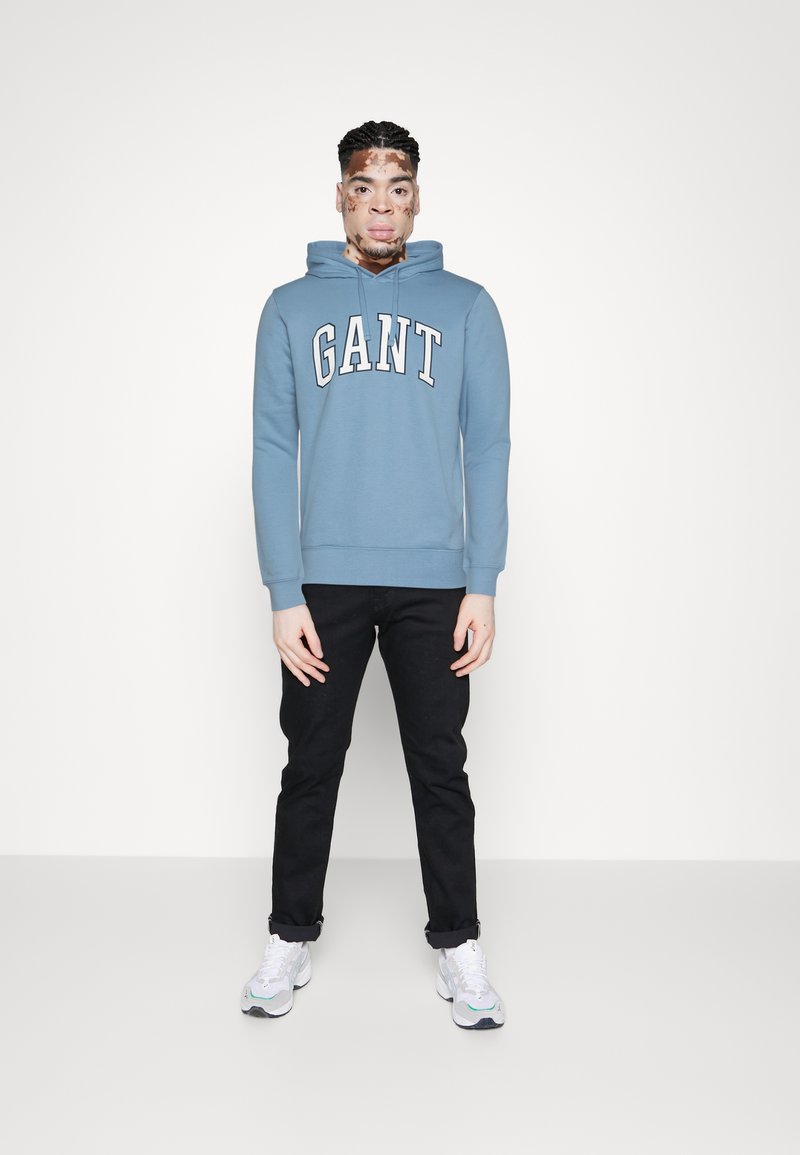 Helle blaue Baumwollkapuzenjacke mit weißem "GANT"-Logo, kombiniert mit schwarzen Hosen und weißen Sneakers. Das Modell steht vor einem einfarbigen Hintergrund.