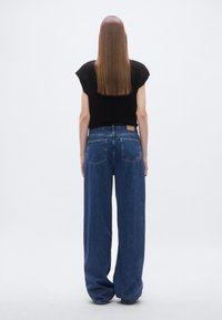 Tamsiai mėlynos spalvos plačios kojų pločio džinsai su aukštu liemeniu; turi nugaros kišenes ir rudos odos prekės ženklo lipduką ant liemens juostos.