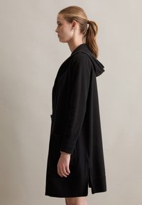 Schwarzer, langer Cardigan mit Kapuze, verfügt über Fronttaschen, gerippte Bündchen und einen seitlichen Schlitz für Bewegungsfreiheit, hergestellt aus weichem, strukturiertem Stoff.