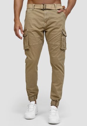 Beige cargobukser med sidelommer, elastiske manchetter, et vævet beige bælte, båret med hvide sneakers på en stående person.