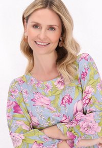 Bluză florală din țesătură albastru deschis, cu modele florale roz și verzi, mâneci gathered și decolteu rotund. Model cu cercei.
