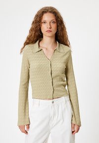 Chemise boutonnée vert clair, texturée, avec col et manches longues, présentant une longueur cropped et un motif de vagues subtil.