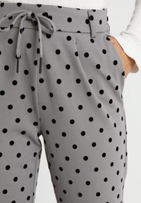Grijze broek met stippen in zwart, voorzien van een trekkoord in de taille en zijzakken. De stof lijkt glad met een lichte glans.